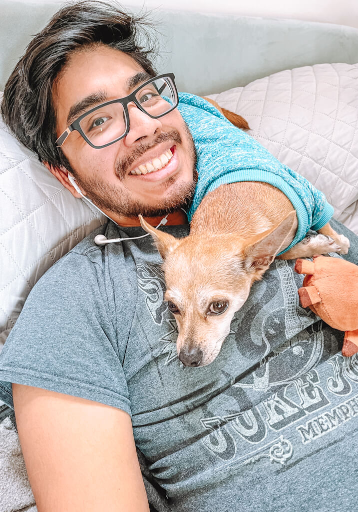 tan chihuahua wearing a blue sweater lying on the shoulder of a man with black hair and glasses on the couch