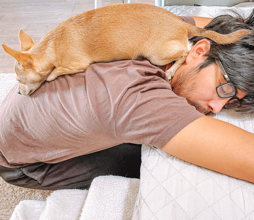 a man wearing glasses and a brown shirt has a dog laying on top of his back, wondering "why is my dog laying on top of me?" 